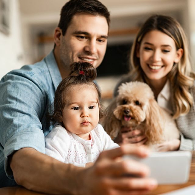 Una familia sonriente compuesta por un hombre, una mujer, una niña y un perro, mirando un dispositivo móvil juntos.
