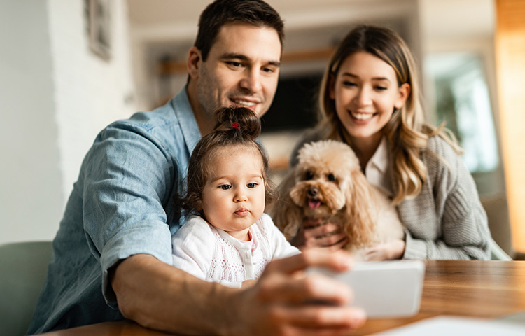 Una familia sonriente compuesta por un hombre, una mujer, una niña y un perro, mirando un dispositivo móvil juntos.