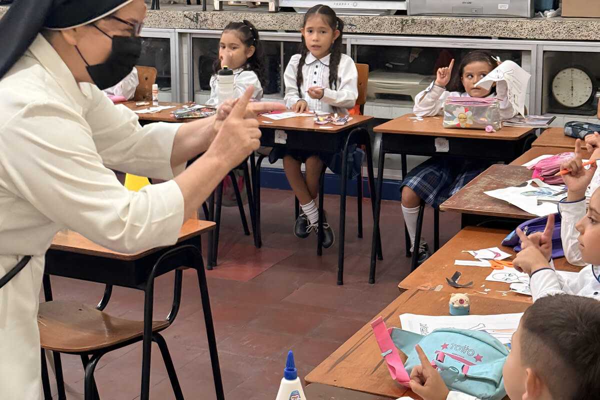 Hermana docente guiando una actividad escolar con niños de primaria, quienes participan levantando la mano y trabajando en manualidades en sus pupitres.