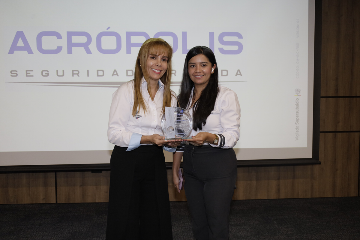 Dos personas sonrientes sostienen un reconocimiento en una ceremonia frente a una pantalla con el logo de Acrópolis Seguridad