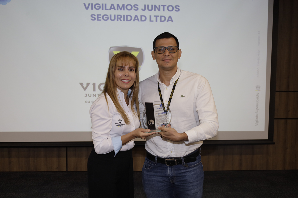 Dos personas sostienen un reconocimiento durante una ceremonia frente a una pantalla con el logo de la empresa Vigilamos Juntos Seguridad LTDA