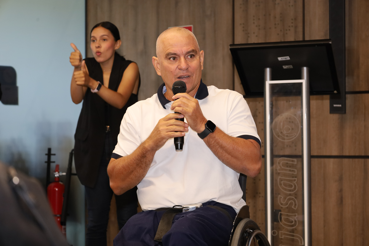 Persona en silla de ruedas hablando en un evento mientras sostiene un micrófono, acompañada de una intérprete de lengua de señas al fondo